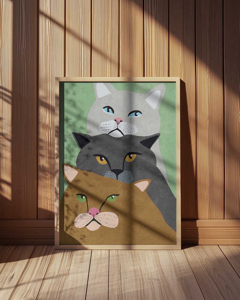 Modern cat art print featuring three minimalist cats in beige, grey, and brown tones with a soft green background. Displayed in a light wooden frame leaning against a wooden wall, illuminated by natural sunlight. Perfect Scandinavian-style wall art for a cozy home.