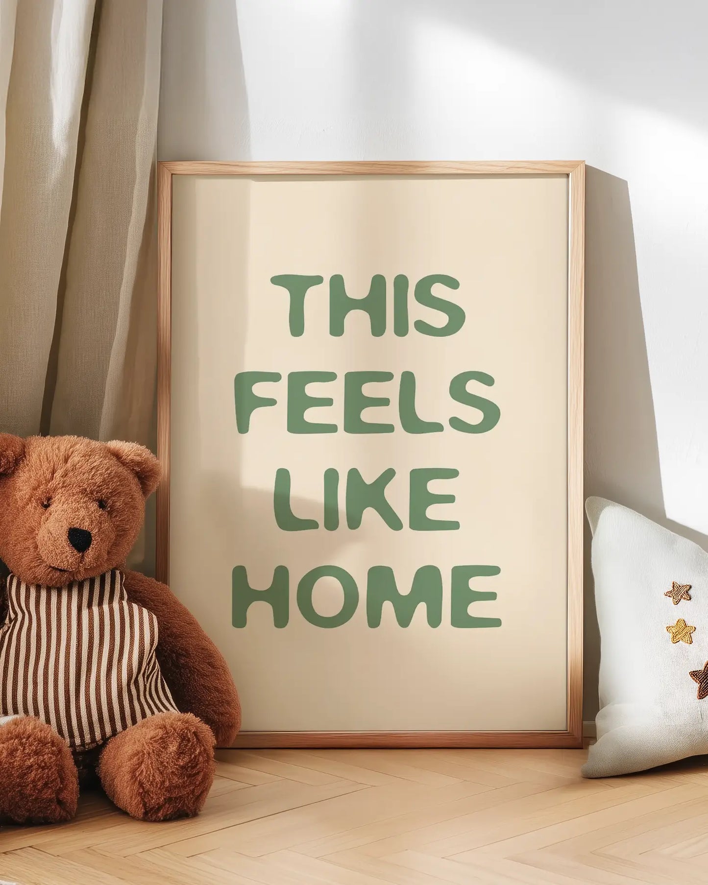 Framed artwork with 'THIS FEELS LIKE HOME' text next to a teddy bear on a wooden floor.